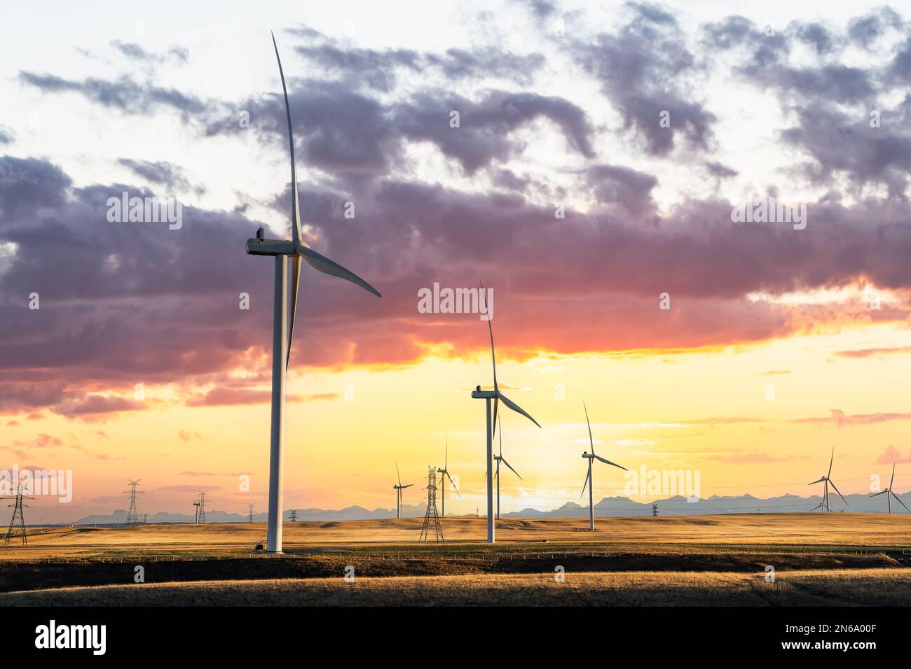Windmills at sunset producing green energy overlooking agriculture wheat fields on a prairie landscape with distant mountains under a dramatic colourf Stock Photo