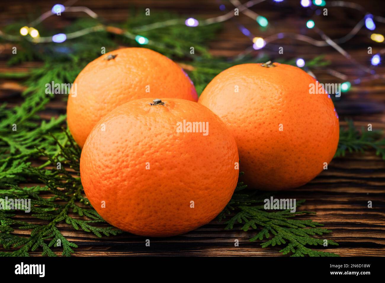 Tangerines close - up on a wooden table near the coniferous branches of juniper on the background of burning lights garlands. Christmas background. Ch Stock Photo