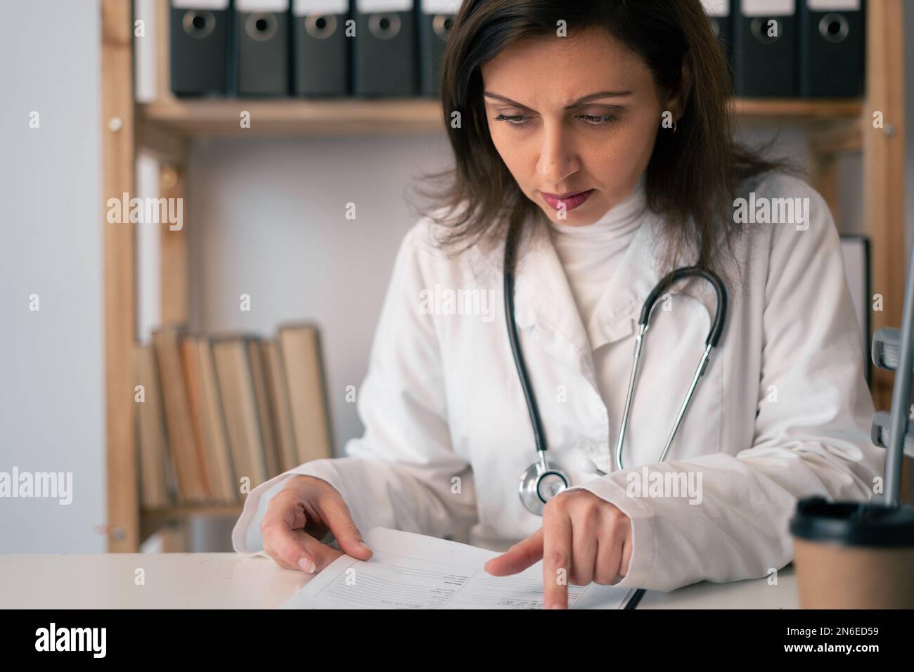 Concentrated female doctor reading document at table in clinic. Doctor
