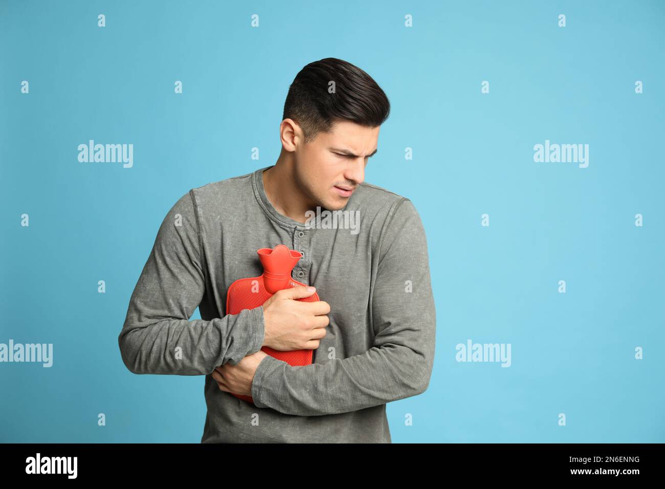 Man using hot water bottle to relieve chest pain on light blue background Stock Photo