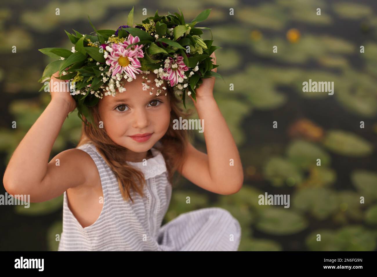 Cute little girl wearing wreath made of beautiful flowers near pond ...
