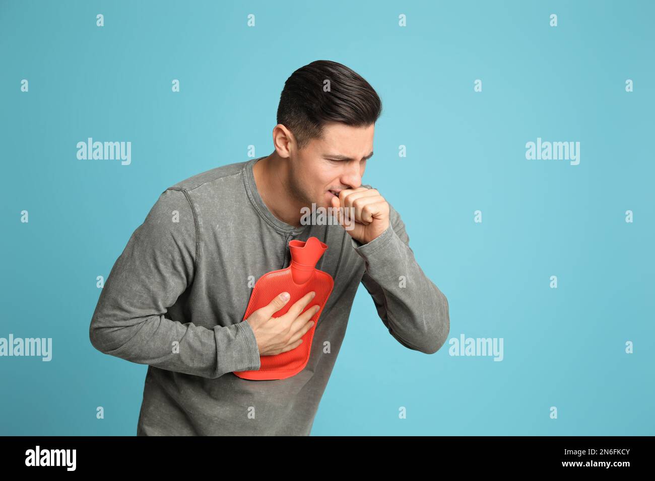 Ill man with hot water bottle coughing on light blue background Stock