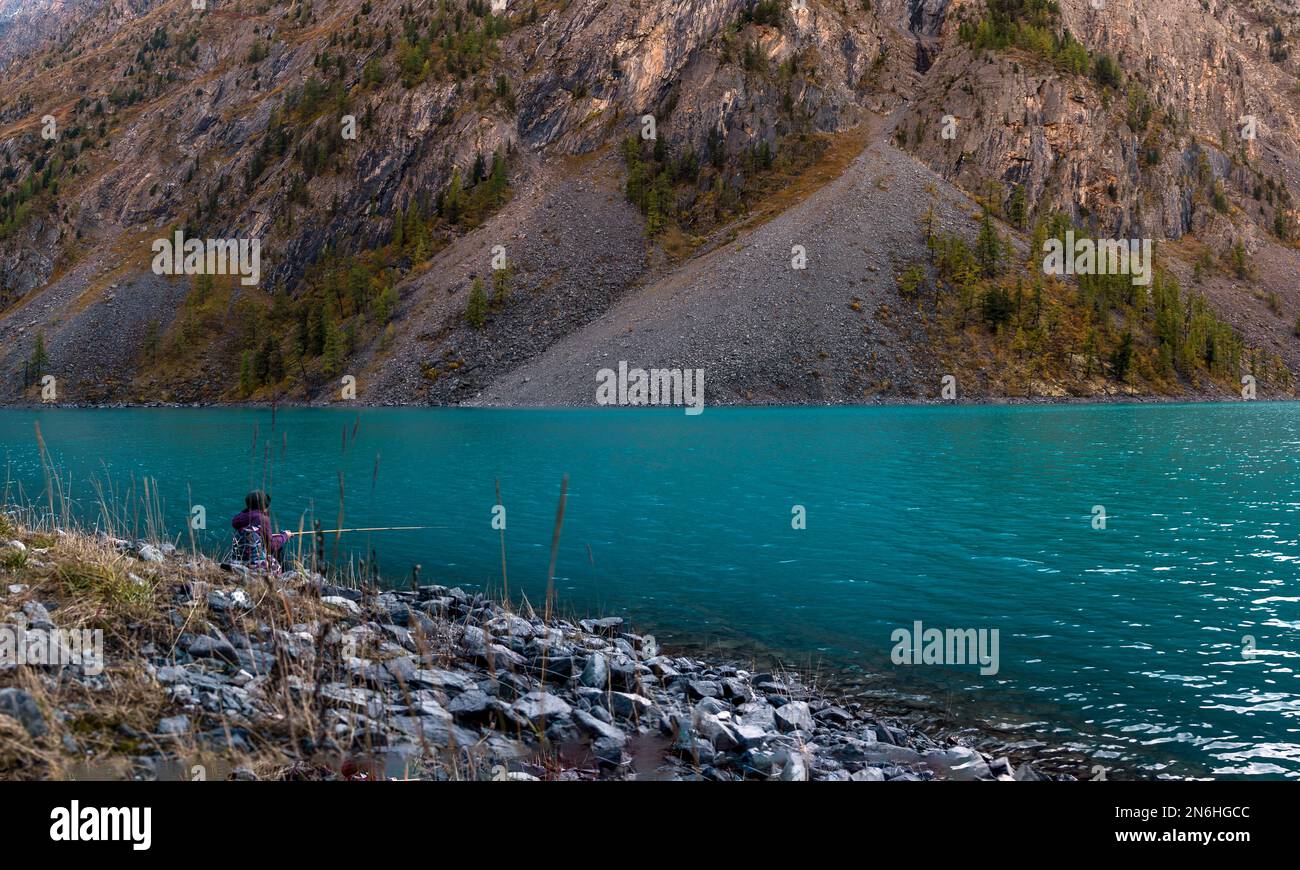 A girl fisherman sits on the shore of the alpine lake Shavlinskoye among the stones of the rocks and catches fish with a fishing rod in the Altai Stock Photo