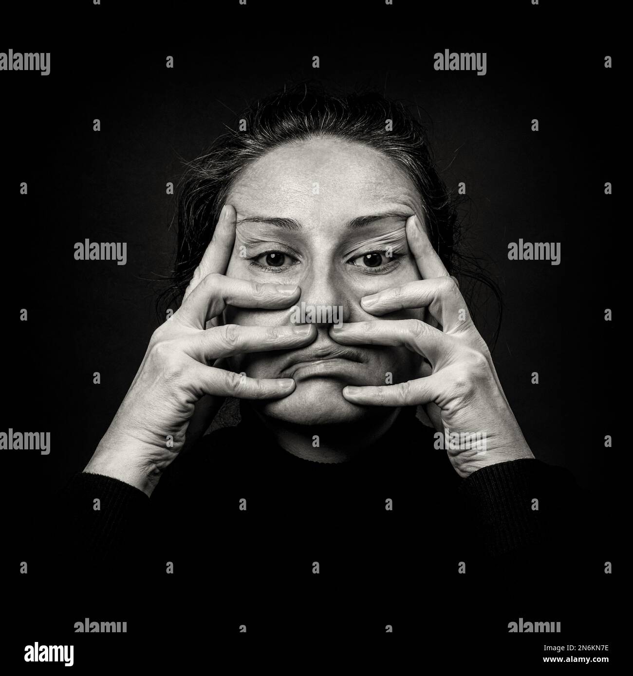 Conceptual dark portrait of old woman stretching the skin of her face in ugly grimace.  Problems with skin and wrinkles, demonstration against plastic Stock Photo