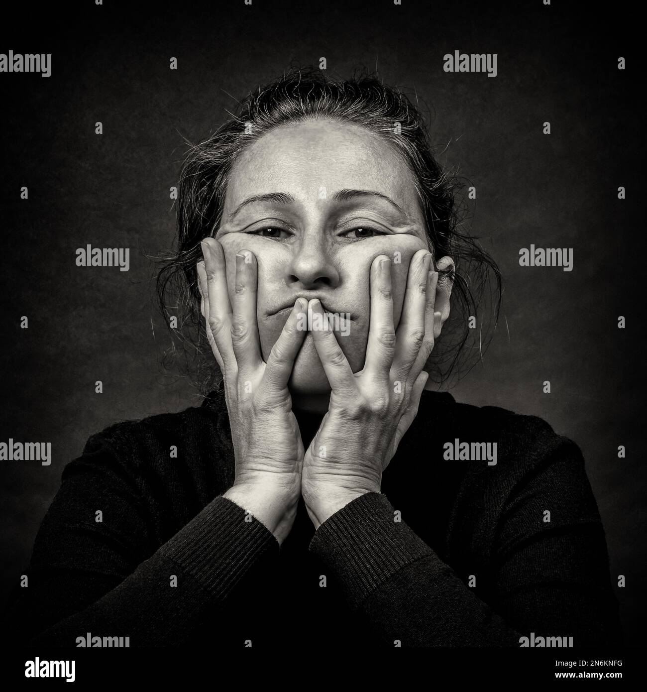 Conceptual dark portrait of old tired woman stretching the skin of her face in ugly grimace.  Black and White film grain. Stock Photo