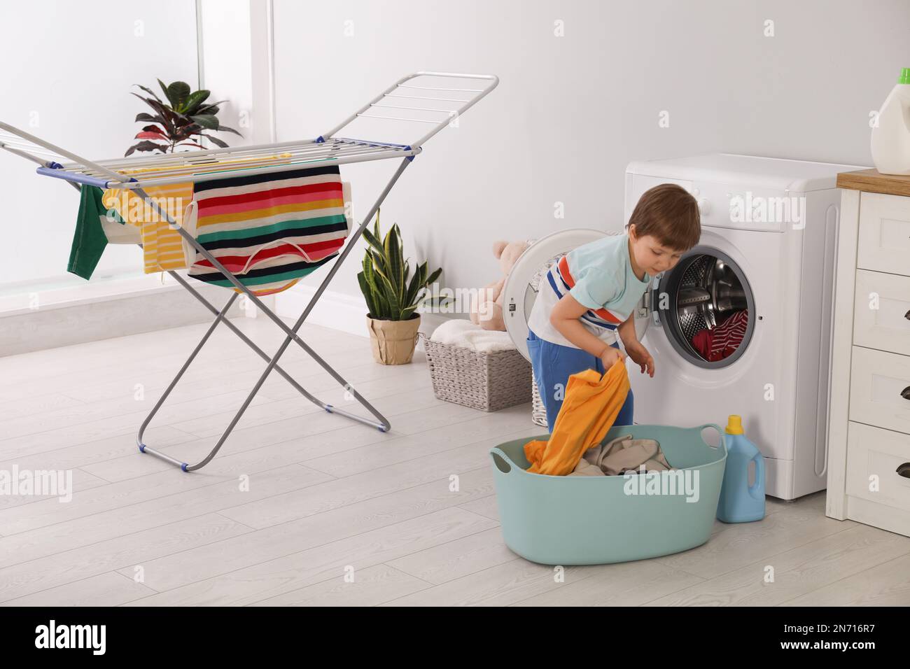 Cute little boy with laundry near washing machine and clothes drying