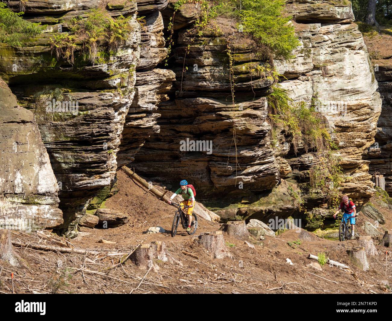 E-mountain biker on single trail in the Mullerthal. Narrow rocky path on the steep slope along the Sauer valley in the direction of Berdorf. For good bikers easy to ride, for beginners borderline, because narrow, partially exposed and littered with stones and boulders. Below flows the border river Sauer, on the other side is the German part of the Müllertal. Stock Photo