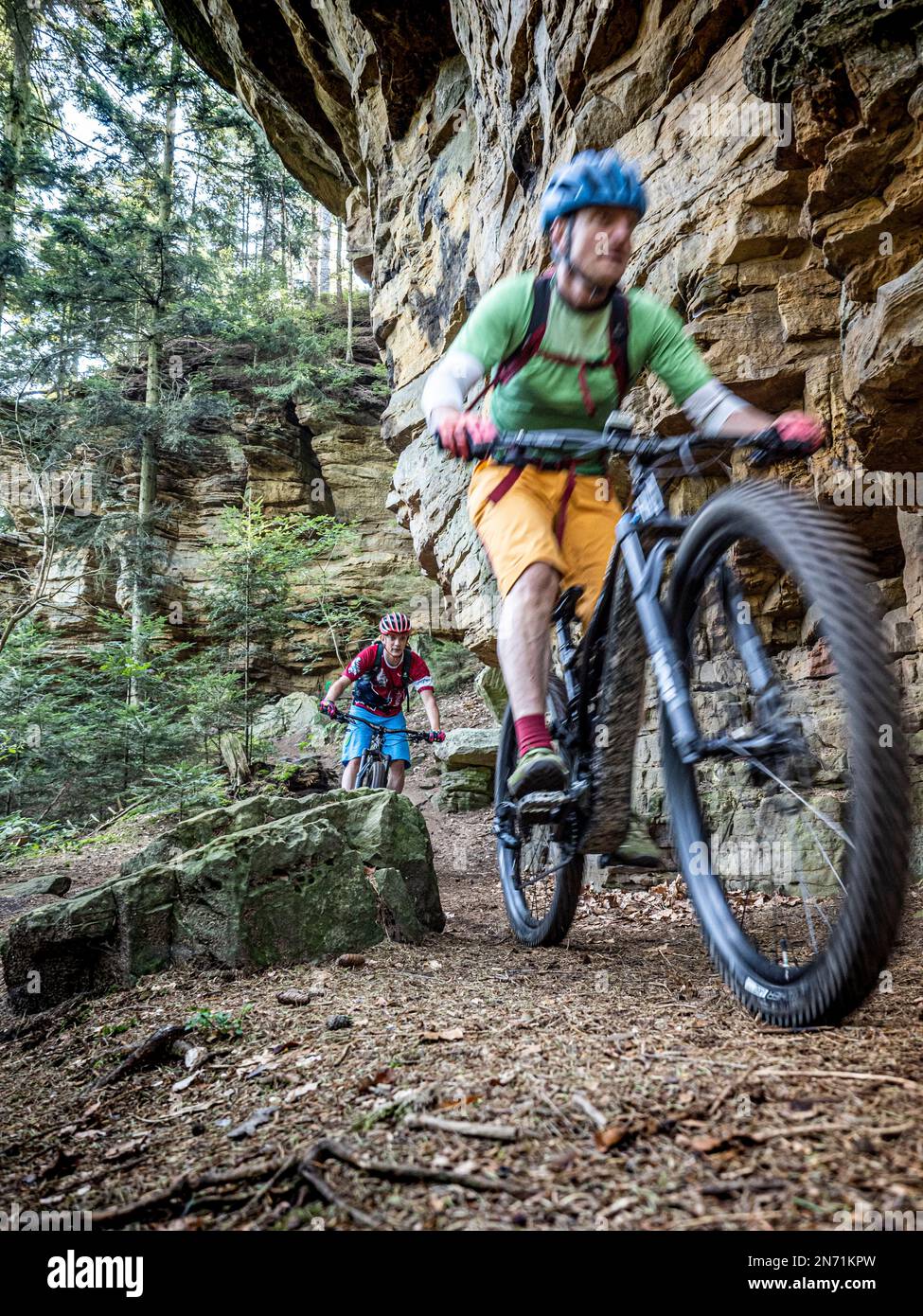 E-mountain biker on single trail in the Mullerthal. Narrow rocky path on the steep slope along the Sauer valley in the direction of Berdorf. For good bikers easy to ride, for beginners borderline, because narrow, partially exposed and littered with stones and boulders. Below flows the border river Sauer, on the other side is the German part of the Müllertal. Stock Photo