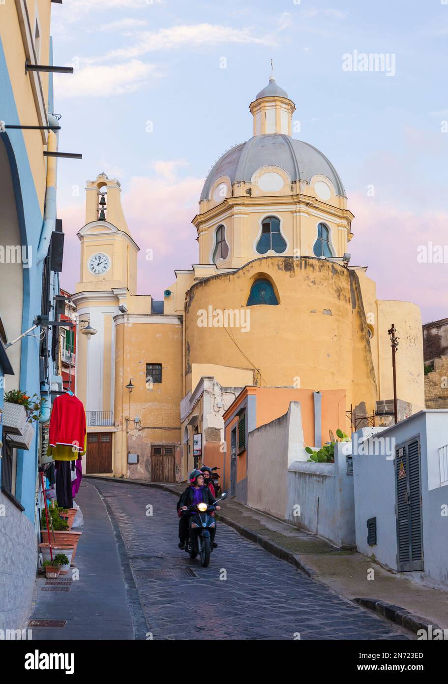 A motor scooter drives down the street San Rocco in the early evening. In the background against pastel sky the church Santiario S. Maria delle Grazie Incoronata. Corricella, Procida, Italy. Stock Photo