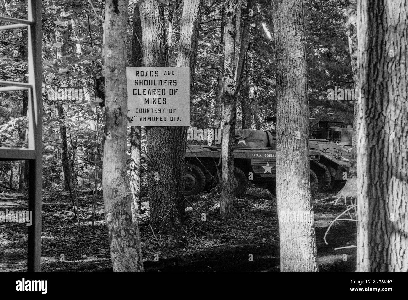 A sign posted on a tree reads Roads and Shoulders Cleared of Mines Courtesy of 2nd Armored Div with a tank parked behind it during a WWII reenactment Stock Photo