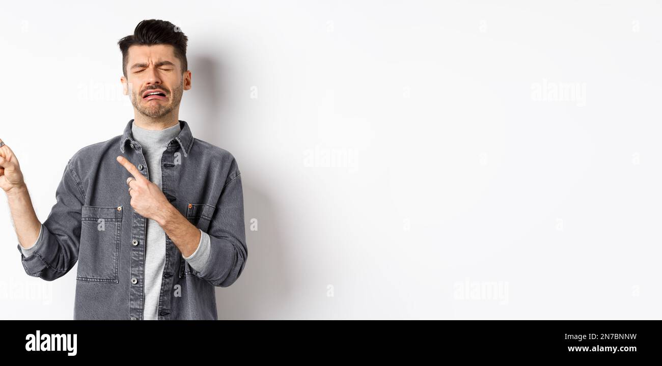 Sad crying man pointing fingers left at logo, sobbing and whining, being jealous or upset, standing on white background Stock Photo