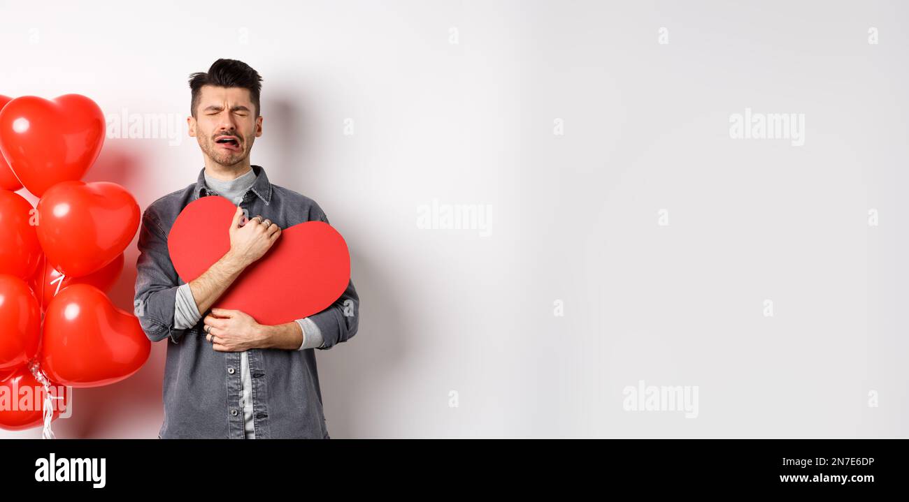 Crying man standing single and lonely on Valentines day, hugging heart cutout and sobbing miserable, being heartbroken and rejected by lover, white Stock Photo