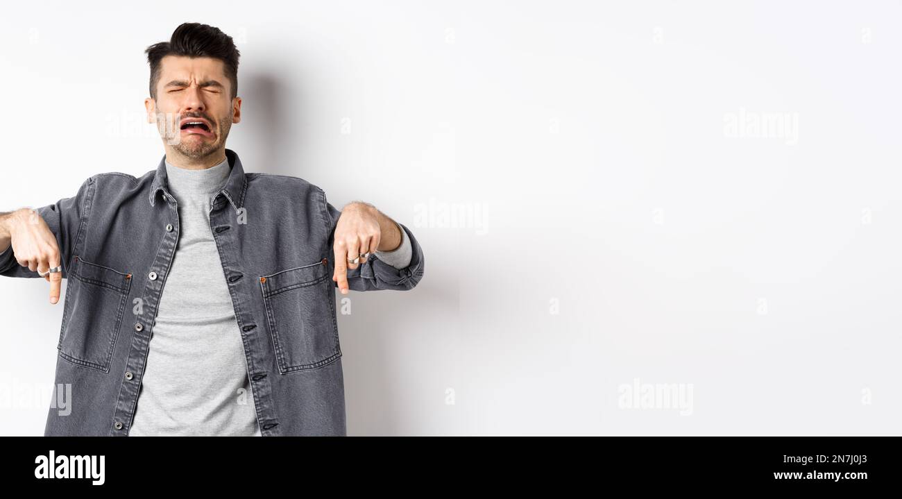 Sad crying man pointing fingers down, sobbing miserable, showing upsetting news, standing in denim jacket on white background Stock Photo