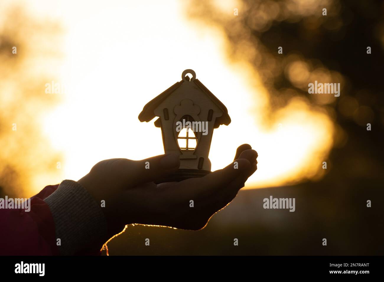 hands hold a small wooden house against the background of the sun at ...