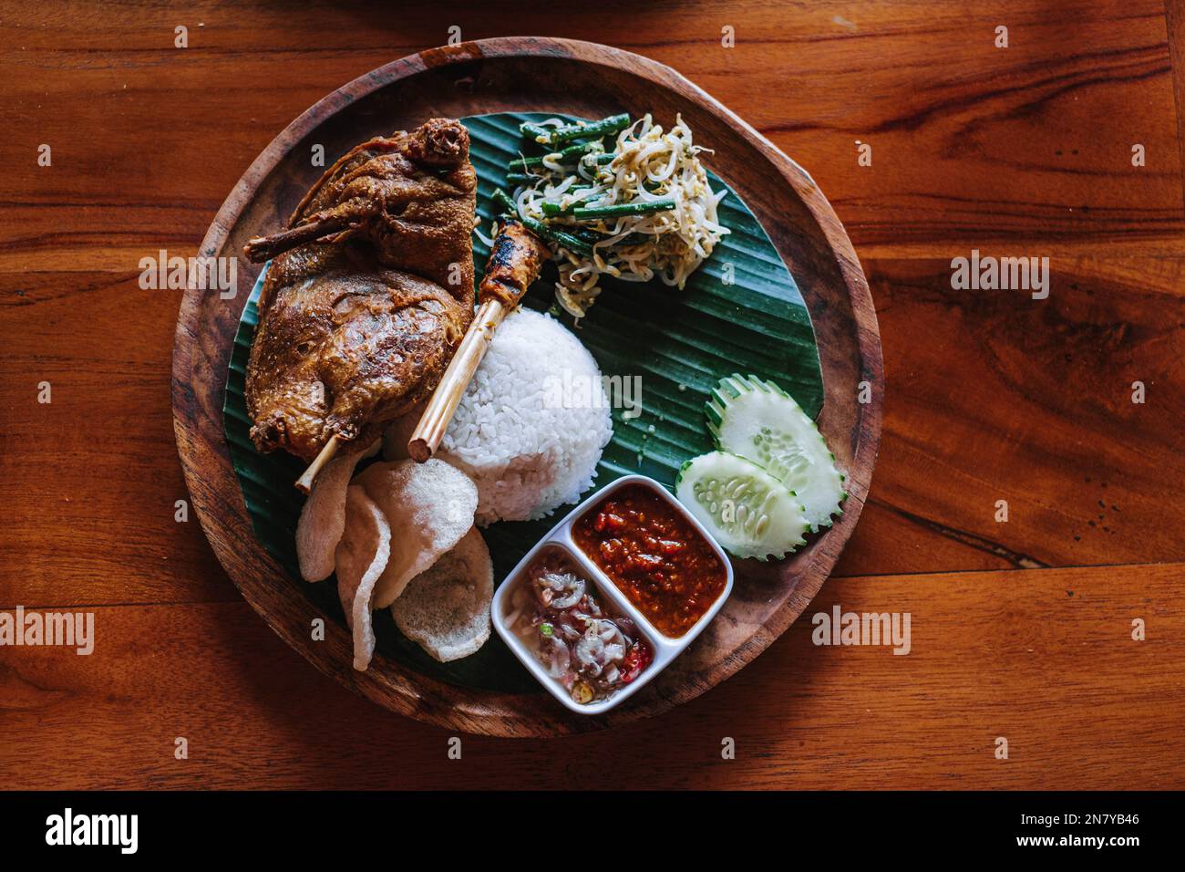 A wooden plate of deep-fried duck with rice, vegetables, chili and crackers. Bebek goreng. Flat lay or top view. Indonesian dish. Stock Photo