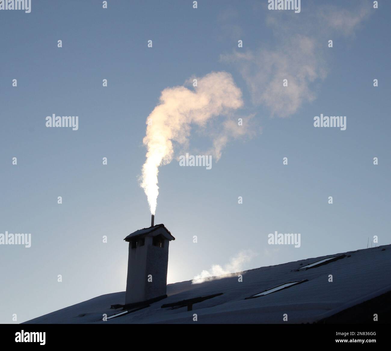 rauchender Kamin im Gegenlicht auf einem verschneiten Hausdach, blauer Himmel * smoking chimney on snowy roof Stock Photo