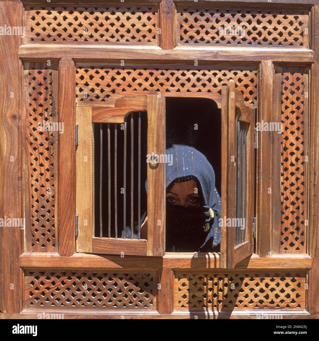 verschleierte Frau mit grossen schwarzen Augen hinter Holzfenster . big eyes of a woman with face veil looking out of a wooden window Stock Photo