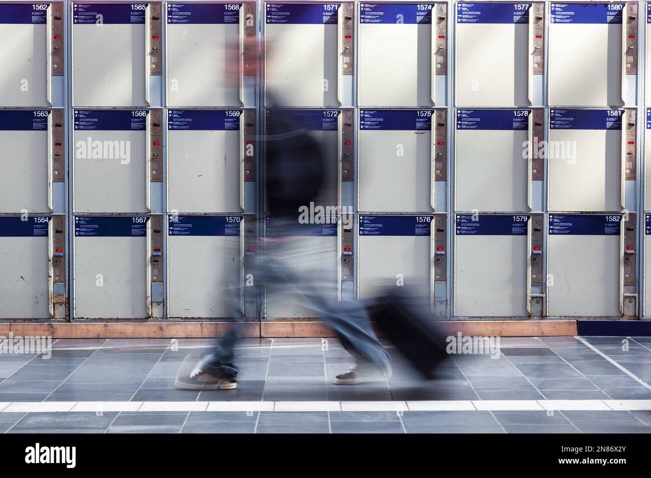 blurred person with a wheeled suitcase walks in front of left luggage lockers Stock Photo