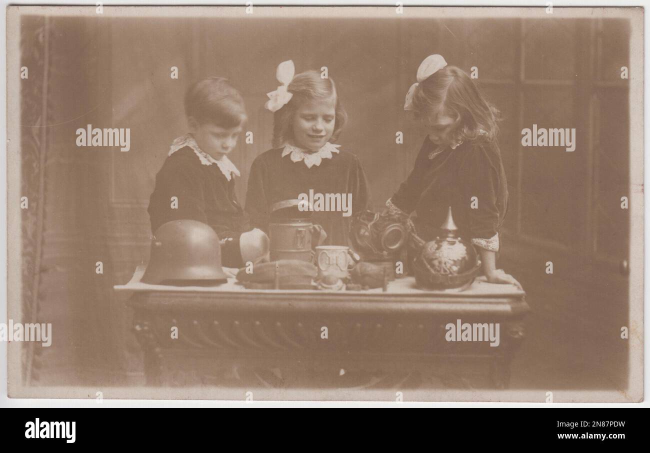 Three British young children looking at First World War trophies taken from German soldiers. The objects laid out on the table include a German Pickelhaube helmet, a Stahlhelm helmet, a field cap, a gas mask and a tin cup or mug. The photograph was taken by the Tasma Studios, 99 Wellington Street, Woolwich, London Stock Photo