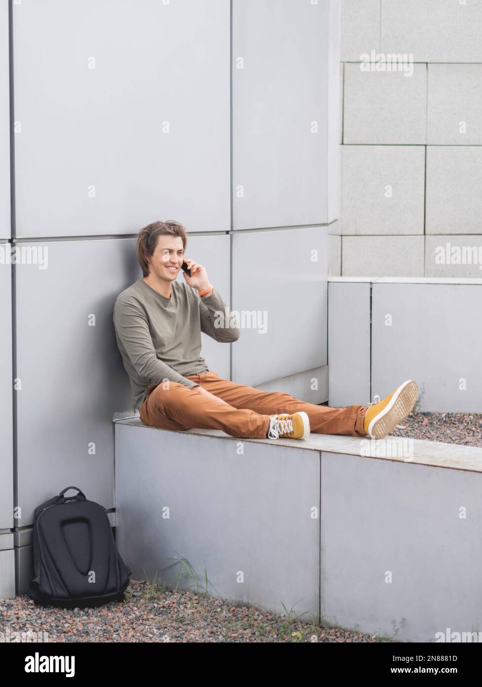 Smiling man sits talks by smartphone on outdoor part of wall. Student with rucksack in casual clothes. Handsome man with long hair. Stock Photo