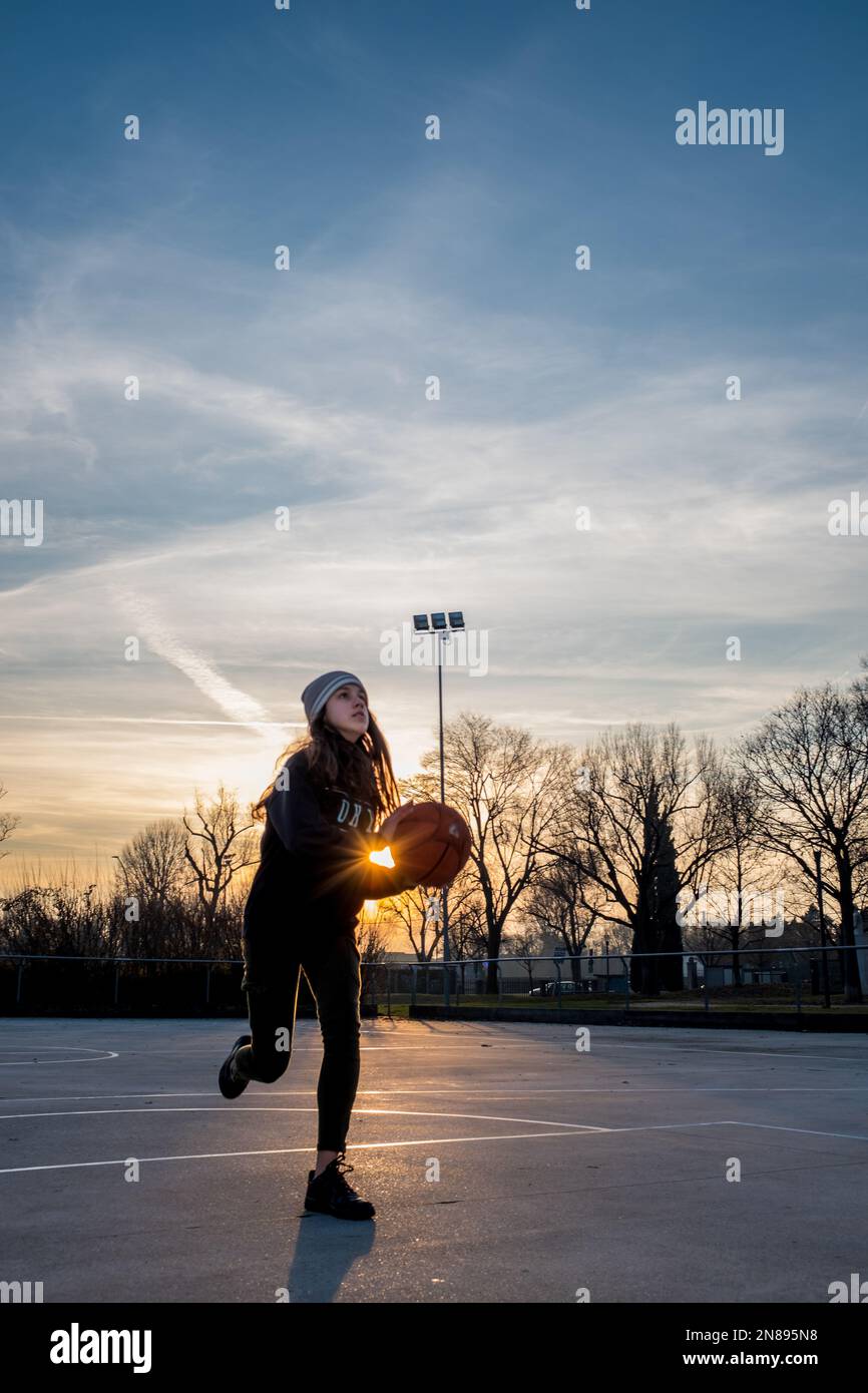 Teenage girl is running with basketball in her hands and looking up in the direction of the basket, alone in an outdoor playground. Stock Photo