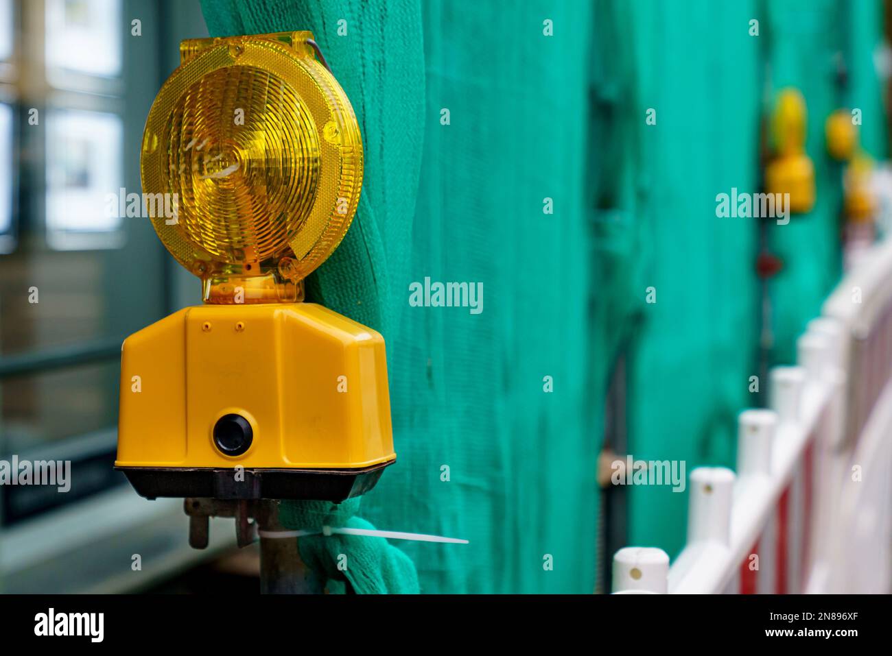 Yellow signal lamp against the background of a green fence of a construction site. Closeup. Stock Photo