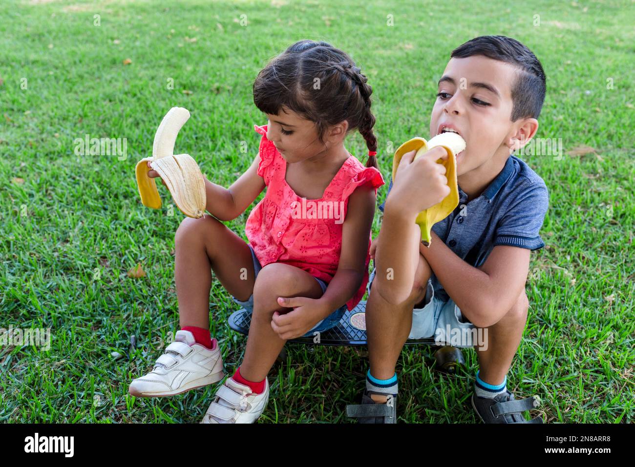 Two kids eating banana sitting outdoors on the grass in a park. Stock Photo