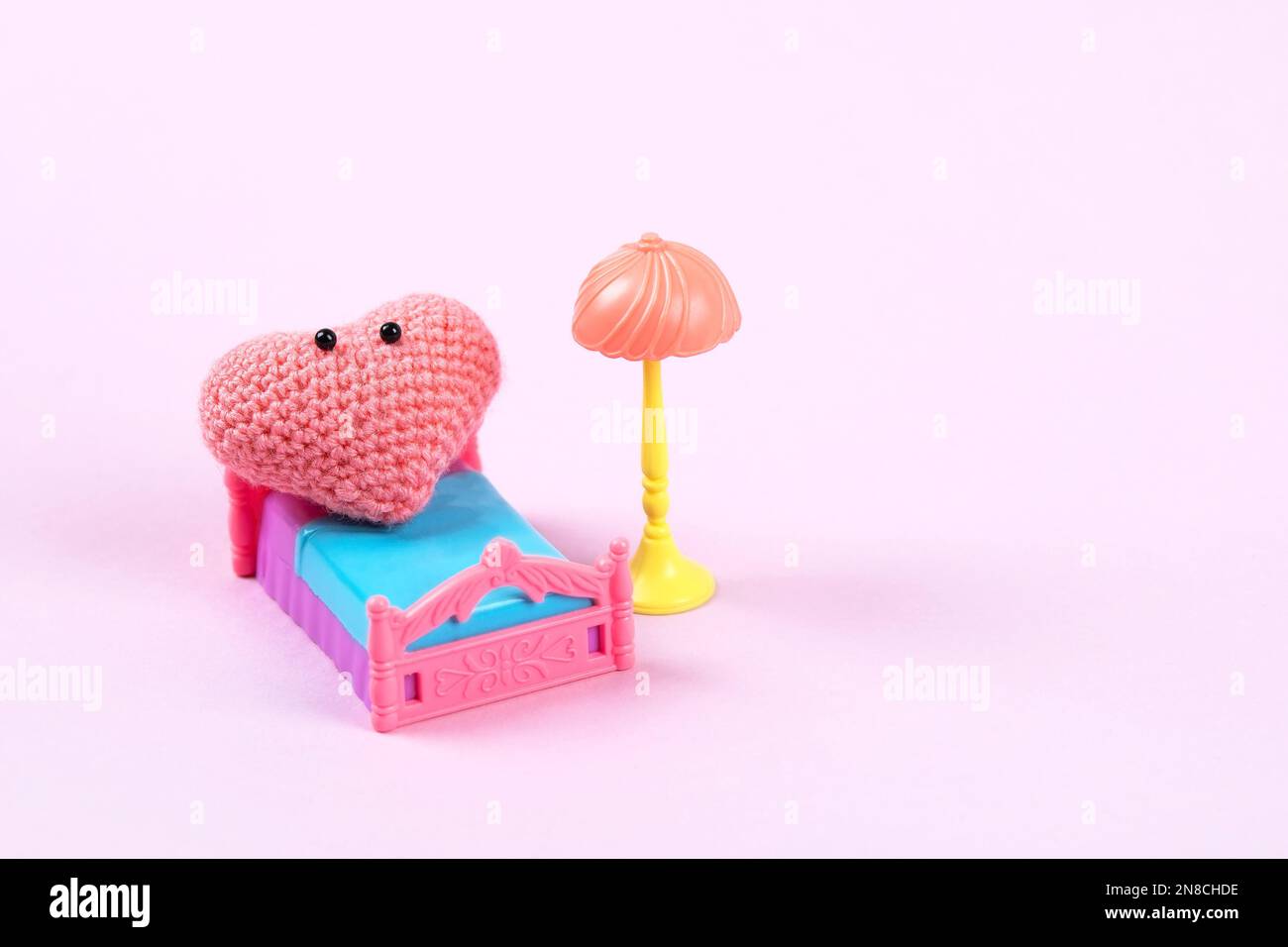 Pink knitted heart with a cute face relaxes on the bed. Love concept. Staged life story of a heart in its room Stock Photo