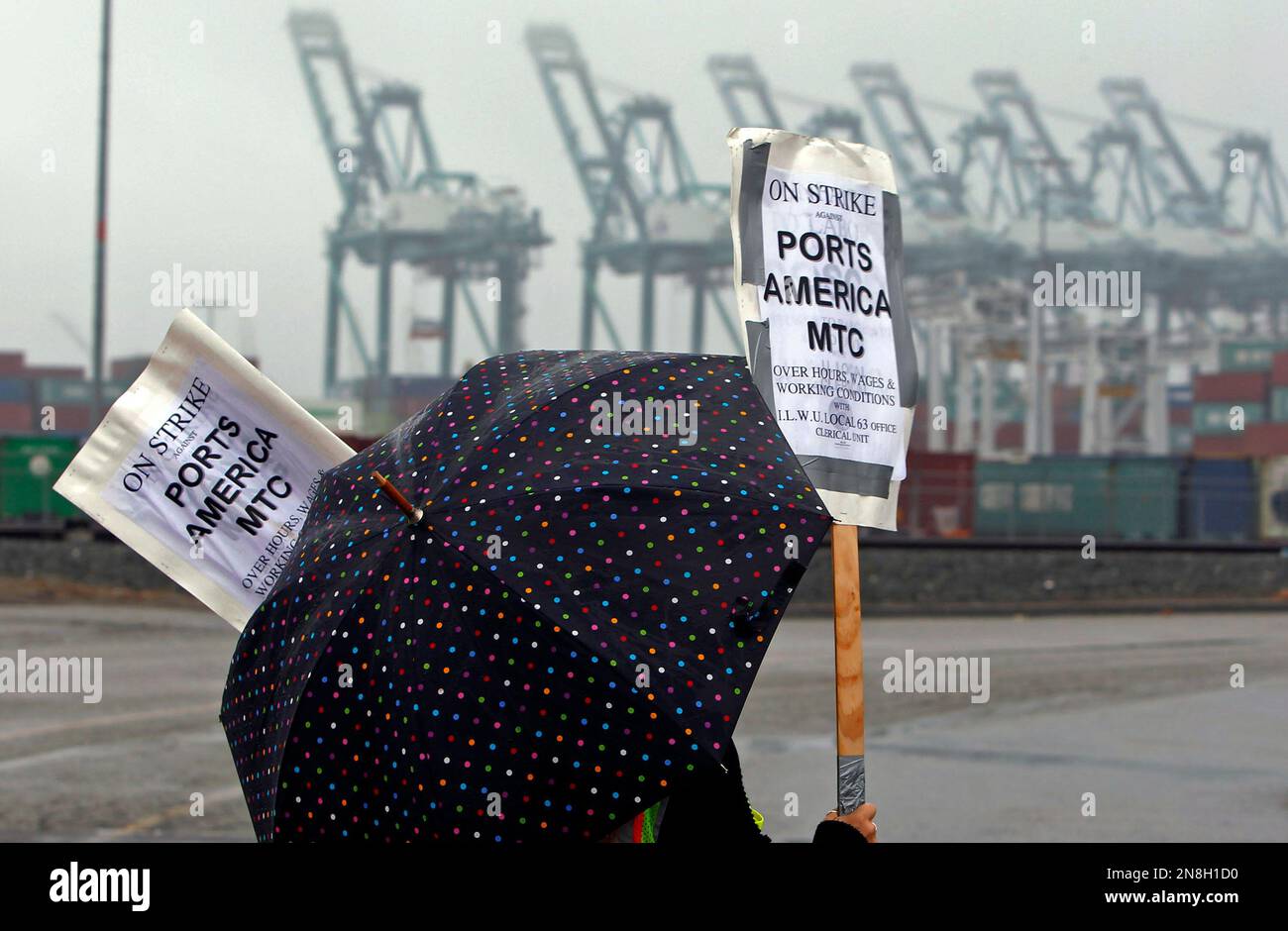 A clerical worker pickets in the rain at the Maersk cargo terminal, where container-handling cranes are in the up and idle position, background, at the Port of Los Angeles Thursday, Nov. 29, 2012. Cargo ships were stacking up at the ports of Los Angeles and Long Beach as a strike by about about 70 clerical workers shut down most of the terminals that together are the nation's busiest port complex. Dockworkers were refusing to cross the picket lines even though an arbitrator ruled the walkout invalid on Tuesday. By Thursday morning, at least 18 ships docked and inside the adjacent harbors were  Stock Photo