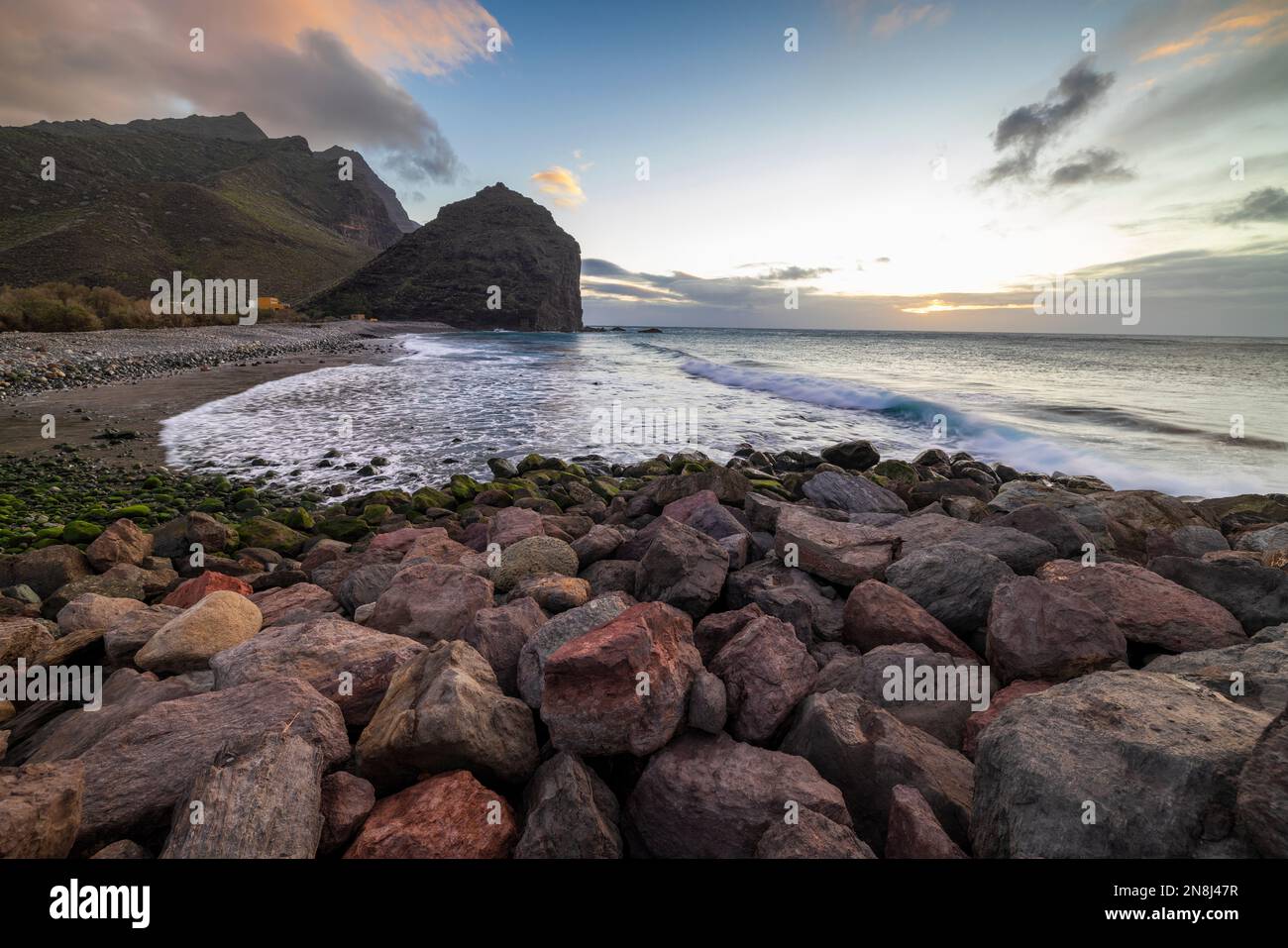 The sunset from the rocky beach. Mountain on the left, ocean on the right, and rocky beach with green stones on the bottom. No people. Some clouds. Stock Photo