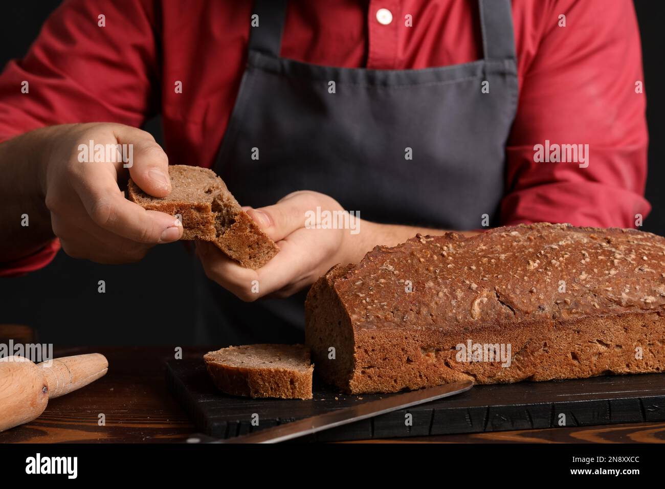 Male baker with slice of rye bread at table on dark background, closeup ...