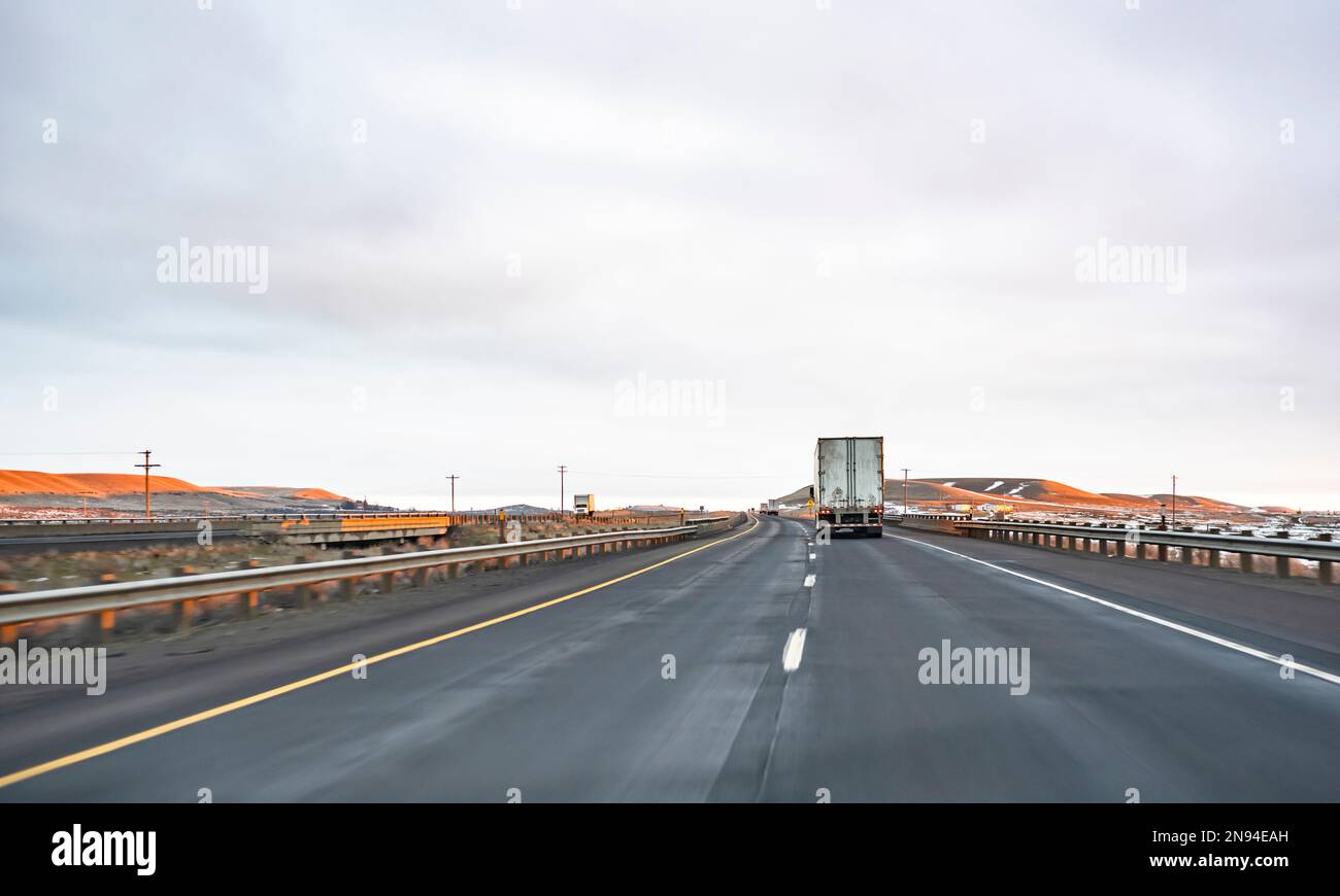 Industrial long hauler big rig semi truck tractor transporting commercial cargo in dirty dry van semi trailer driving on the straight highway road bet Stock Photo