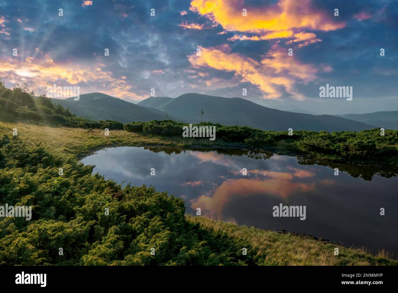 Colorful sunset sky reflecting in the small mountain lake on alpine meadow. Dramatic outdoor landscape. Stock Photo