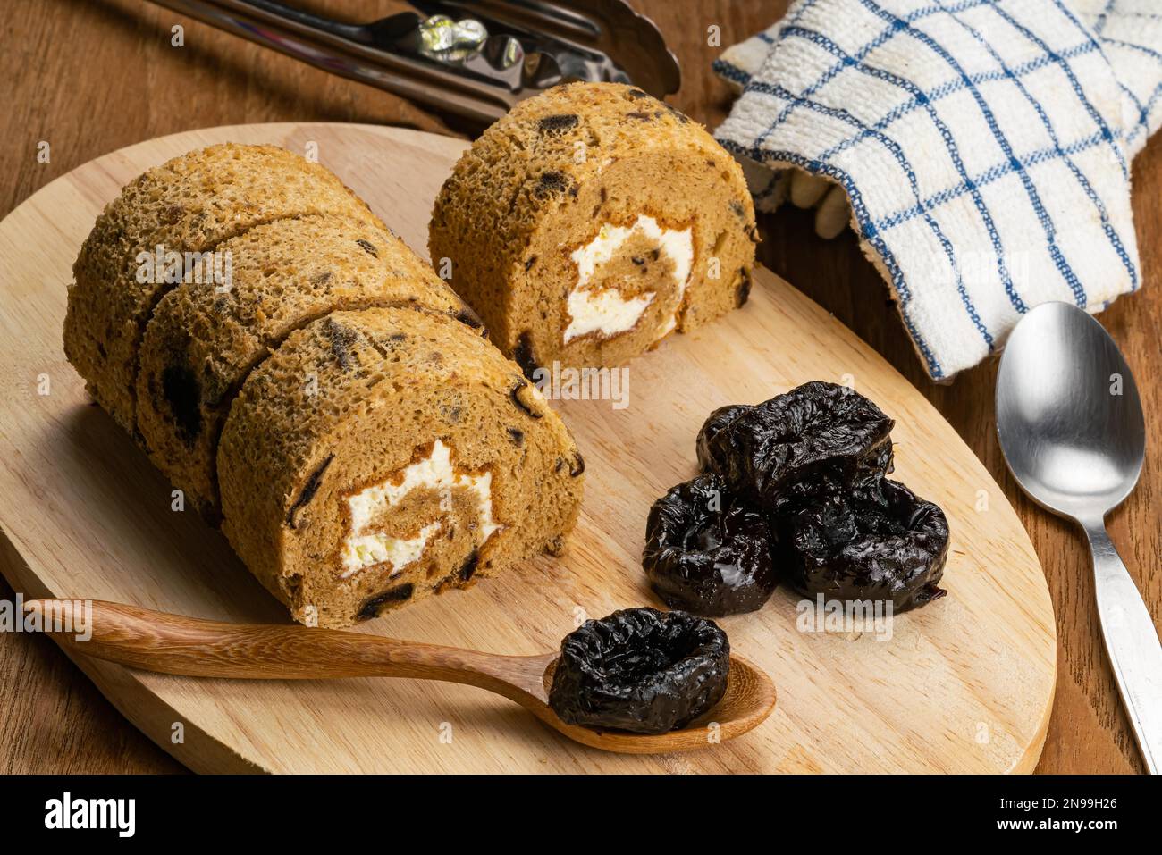 Hogh angle view of homemade sponge cake roll and dried pitted prune fruit on wooden board with metal spoon, metal tongs and tablecloth on wooden table Stock Photo