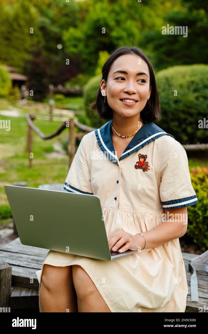 Young cheerful asian woman wearing dress using laptop while sitting on ...