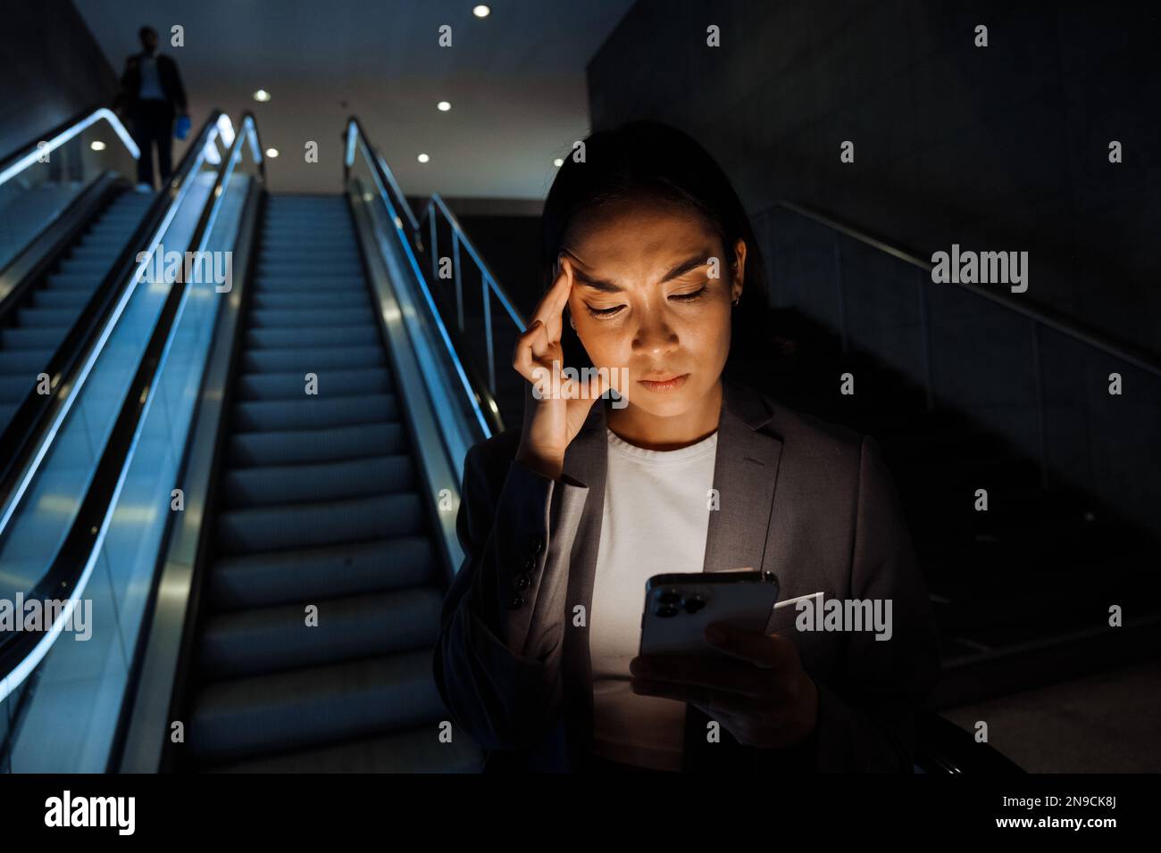 Young displeased asian woman wearing suit frowning while using smartphone standing on escalator indoors Stock Photo