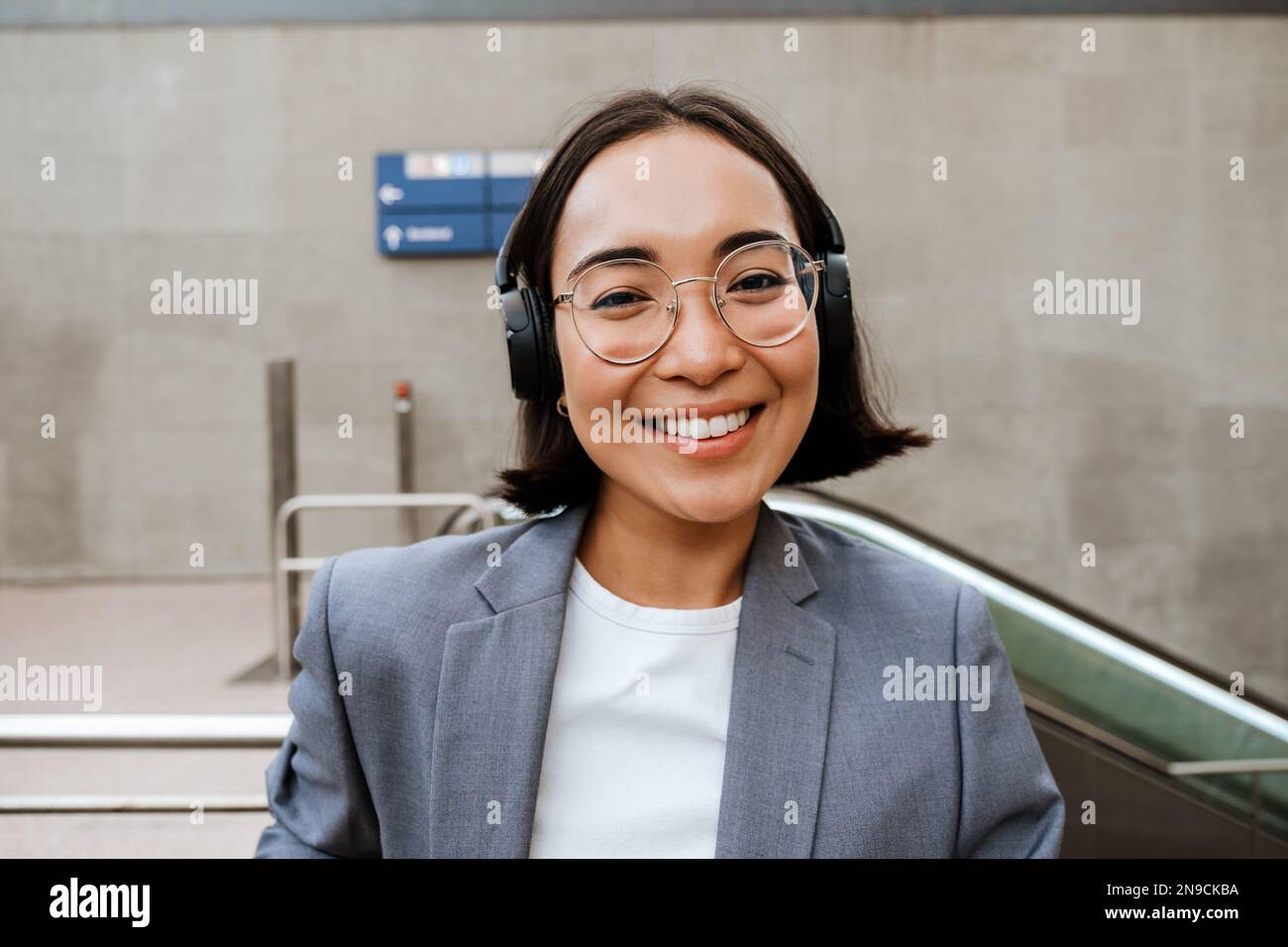 Young asian woman in headphones smiling and looking at camera while standing outdoors near the escalator Stock Photo