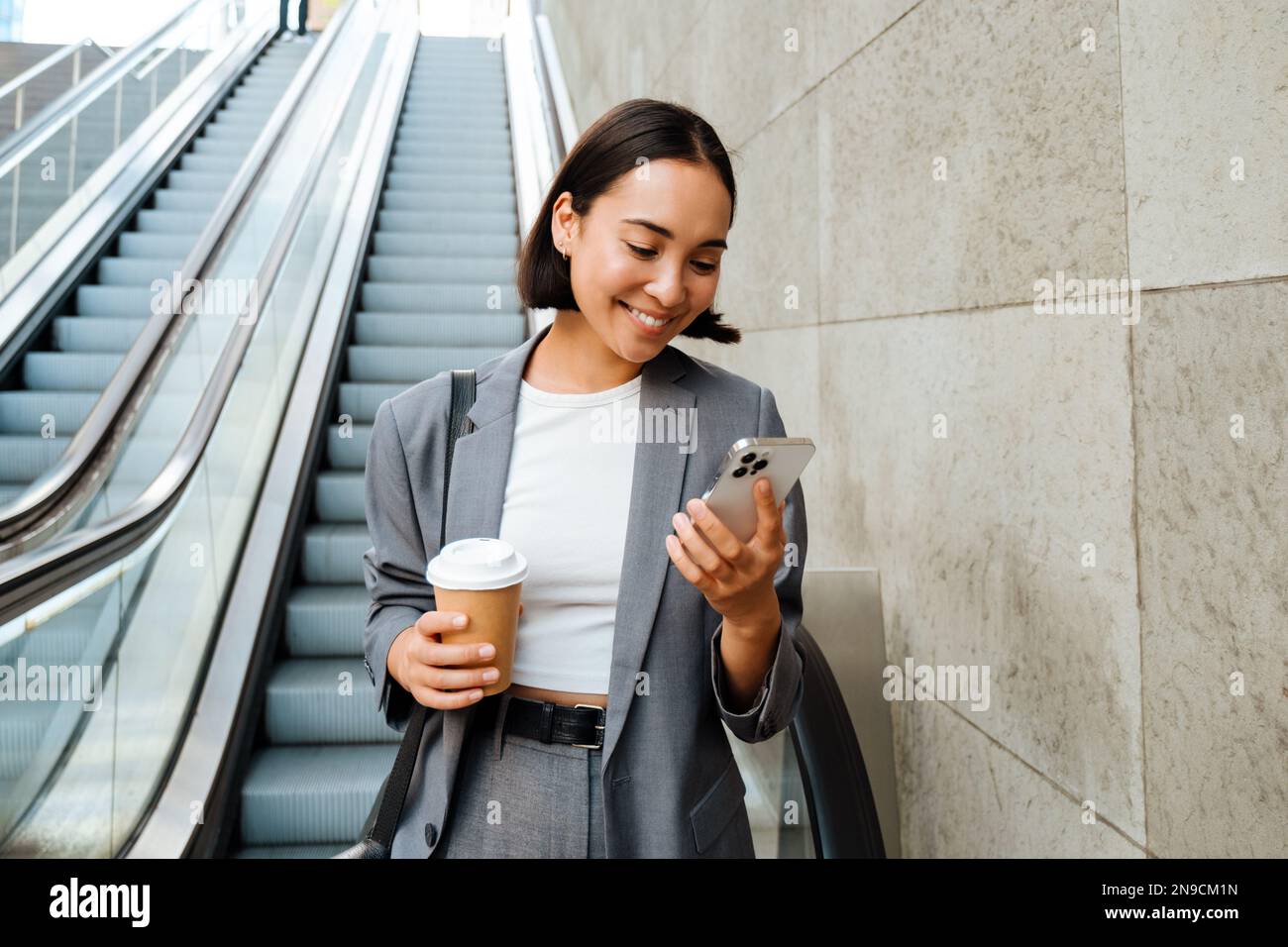 Young smiling asian woman holding coffee and using mobile phone while standing on escalator outdoors Stock Photo