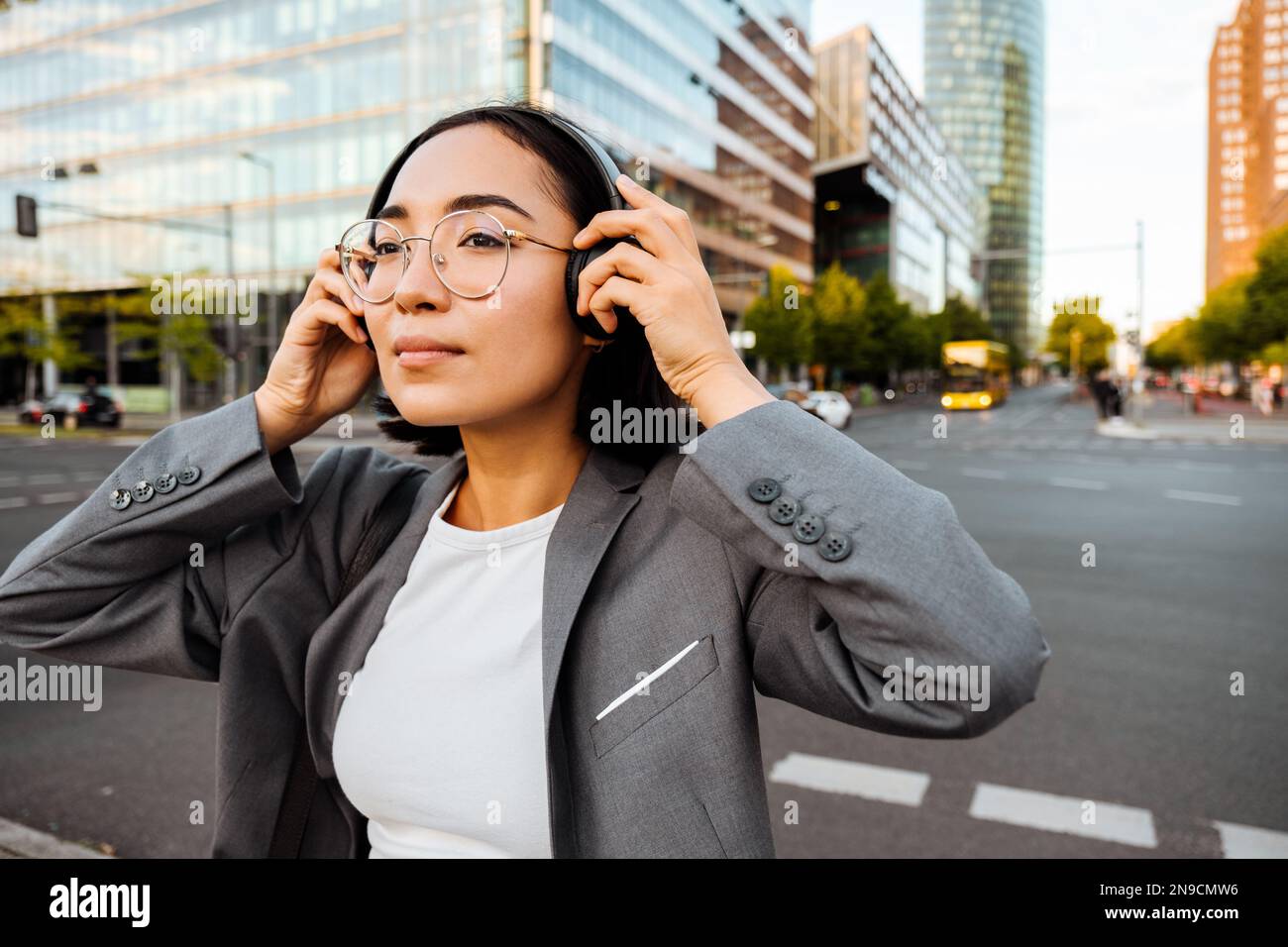 Young pretty asian woman in eyeglasses putting on headphones while standing at city street Stock Photo