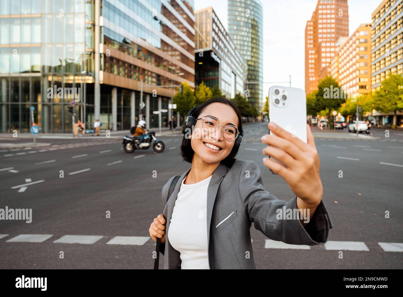 Young joyful asian woman in headphones taking selfie on smartphone while standing at city street Stock Photo