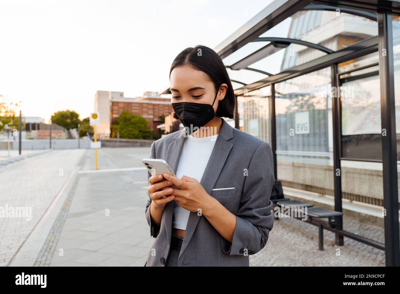 Young asian woman in medical face mask using mobile phone while standing at bus station Stock Photo