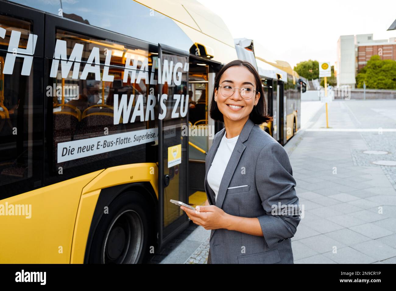 Young asian woman in glasses smiling and holding smart phone while standing near the bus Stock Photo