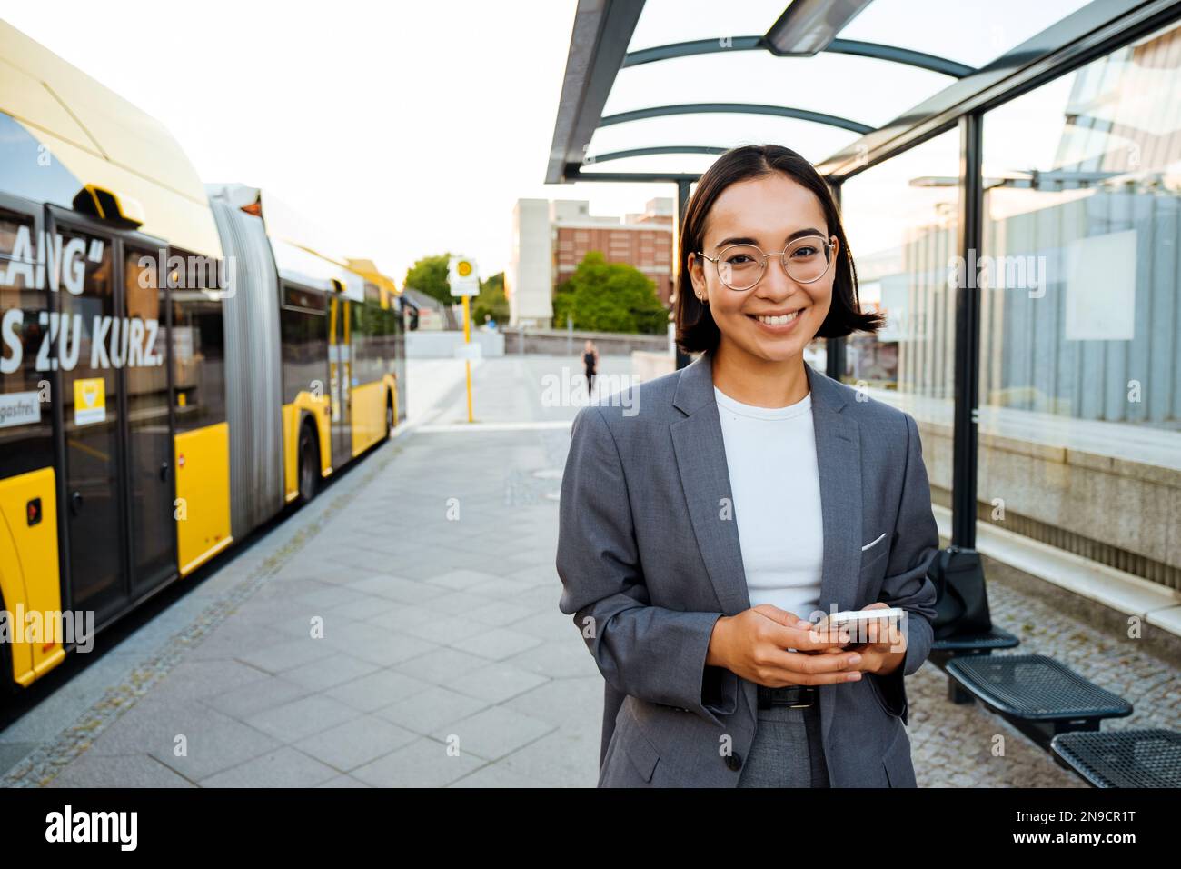 Young asian woman in glasses smiling at the camera and using mobile phone while standing at bus station Stock Photo
