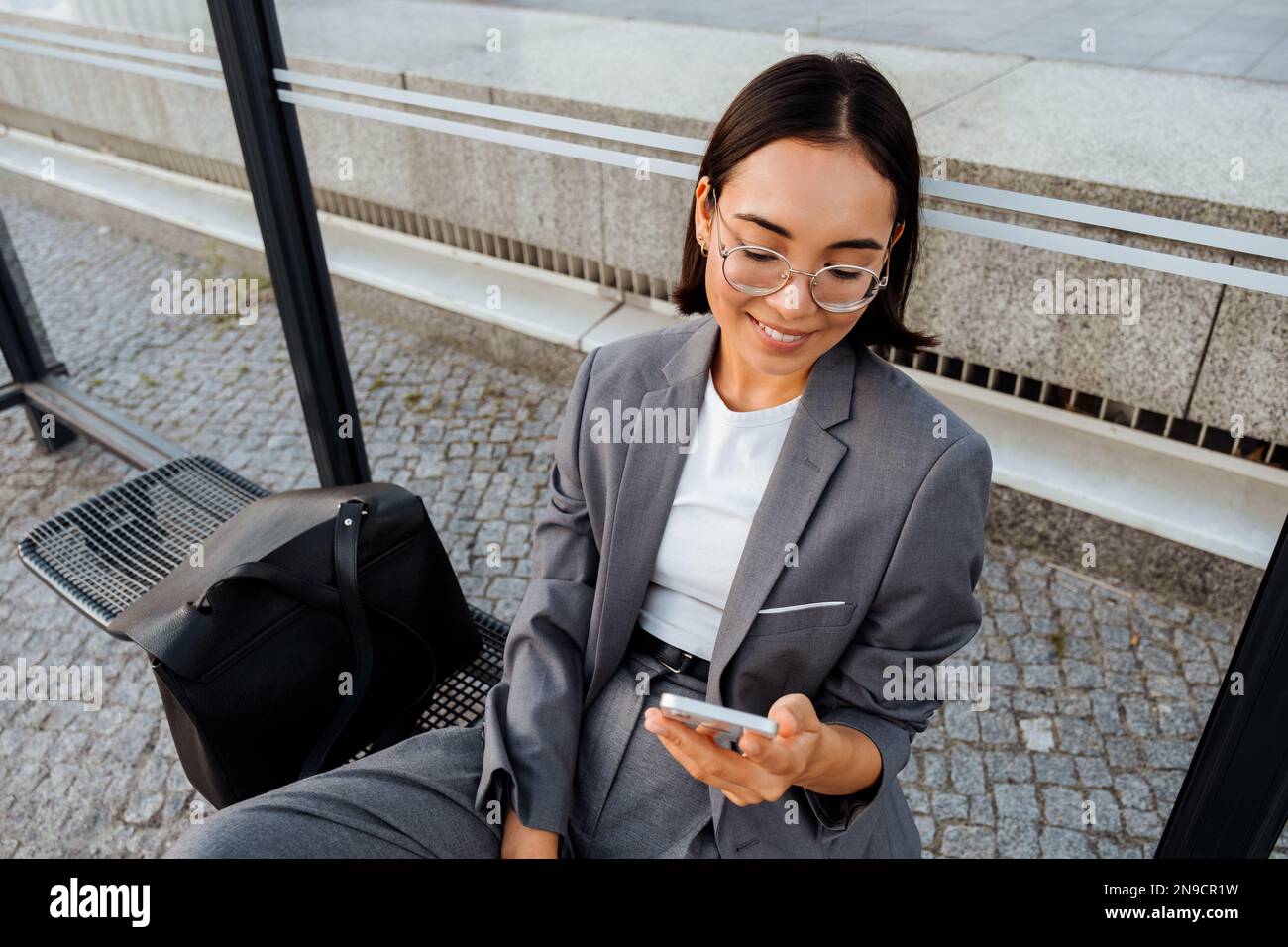 Young asian woman in glasses smiling and using mobile phone while sitting at bus station Stock Photo