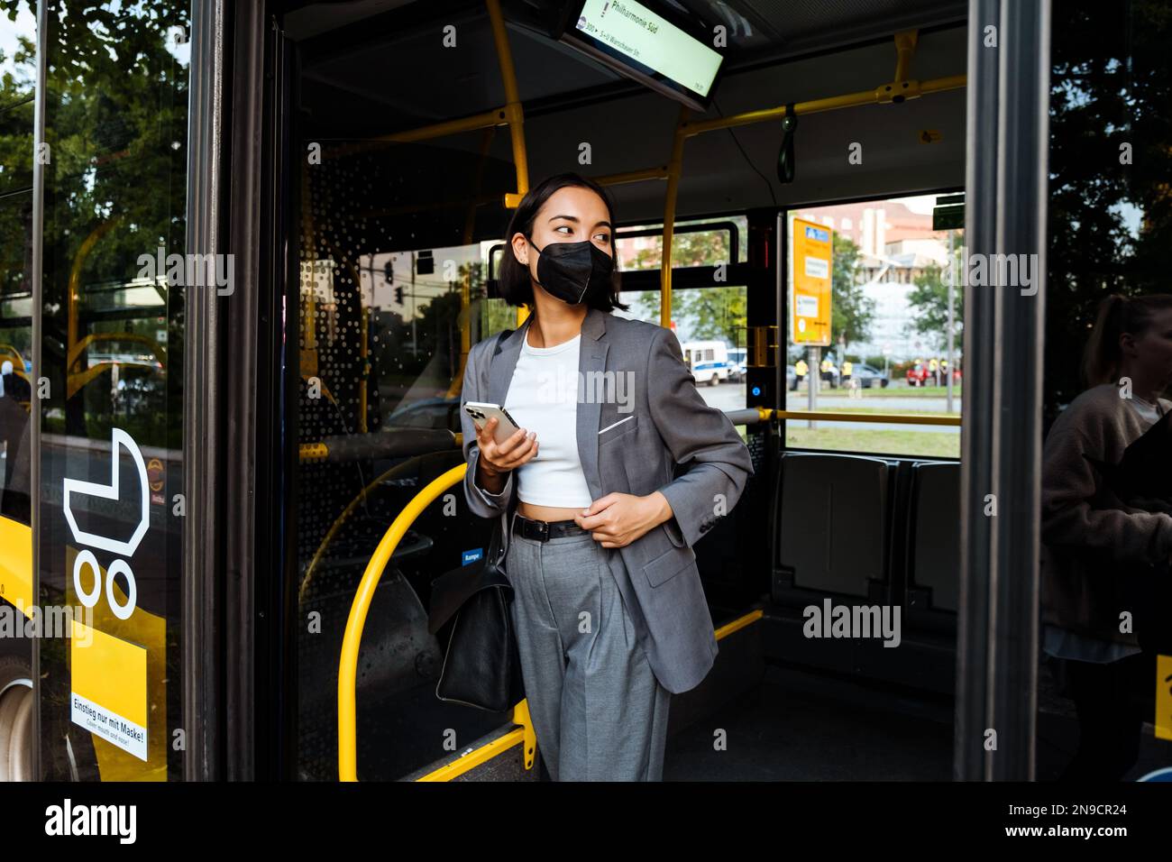Young asian woman in medical mask with a smartphone in her hands looking aside while standing in public bus Stock Photo