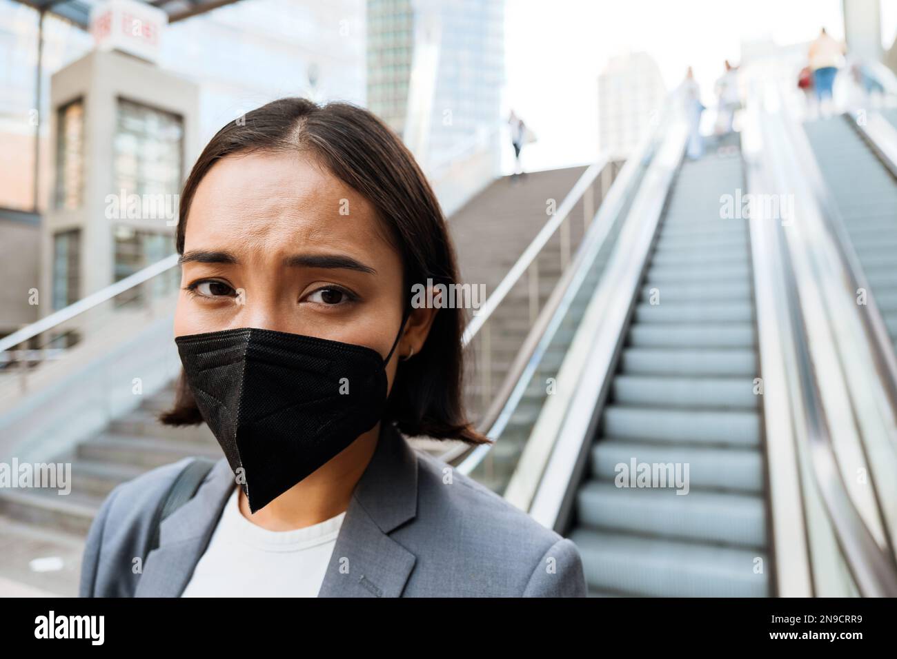 Young asian displeased woman in medical mask standing on escalator outdoors Stock Photo