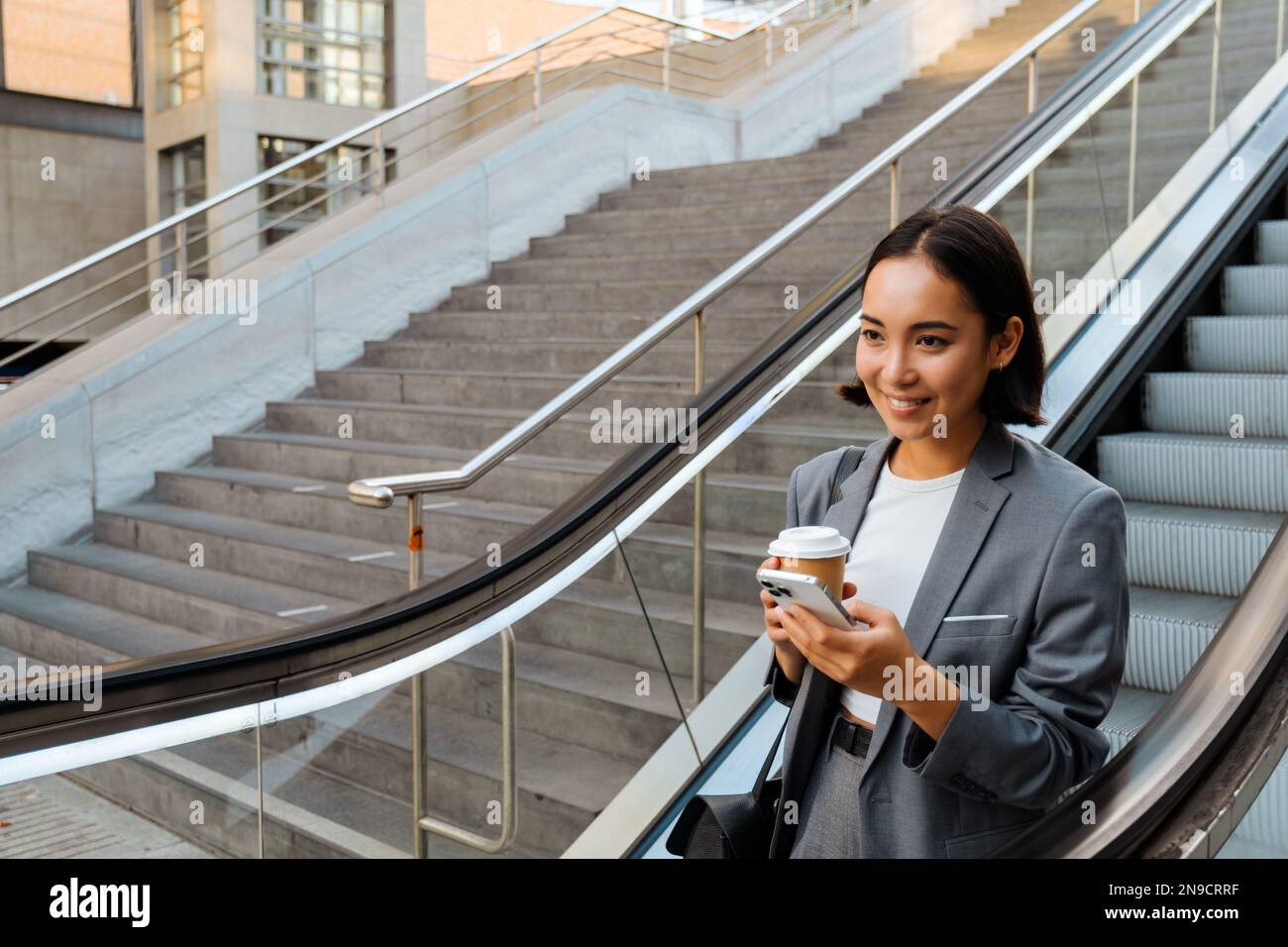 Young asian woman using cellphone and holding coffee while going down escalator outdoors Stock Photo