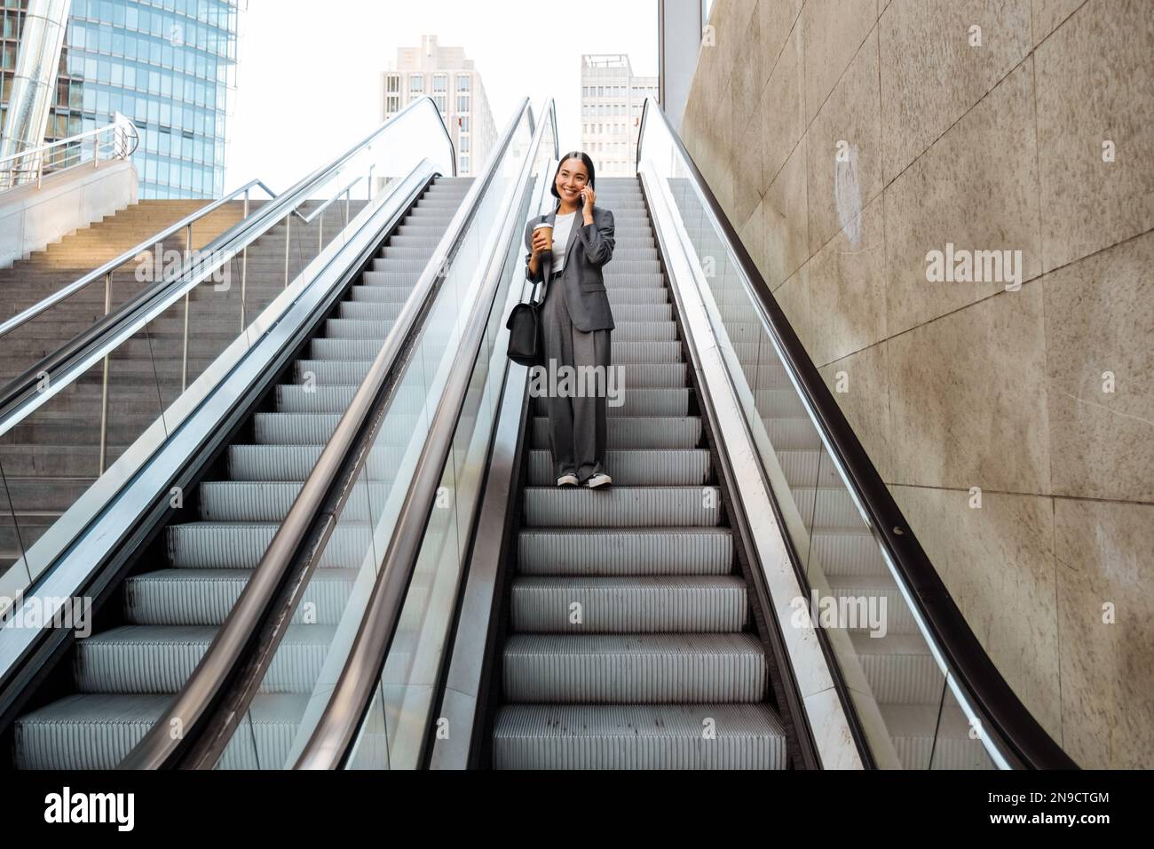 Young asian woman talking on cellphone and holding takeaway coffee while going down escalator outdoors Stock Photo