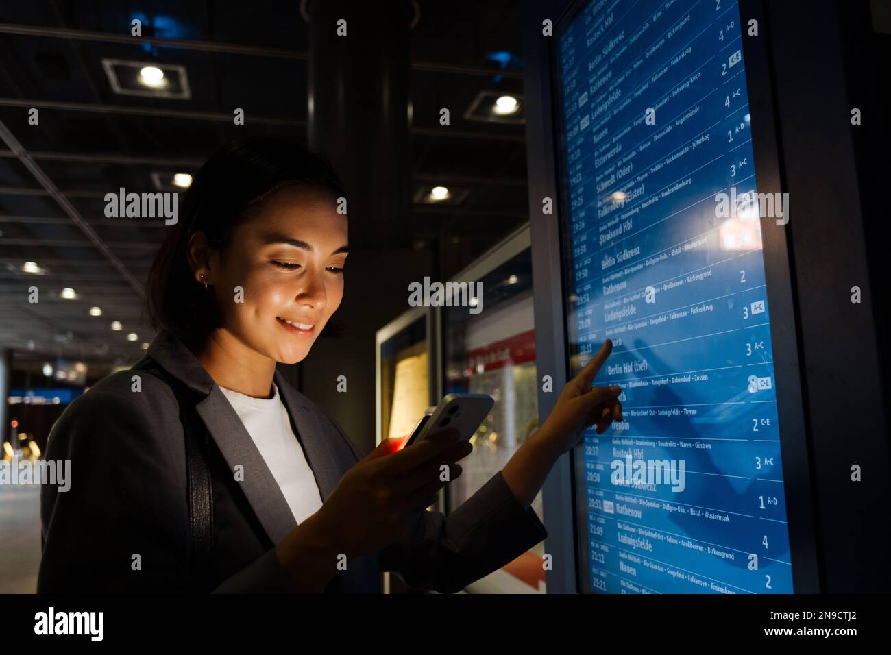 Young asian woman using mobile phone and pointing finger at the electronic timetable indoors Stock Photo