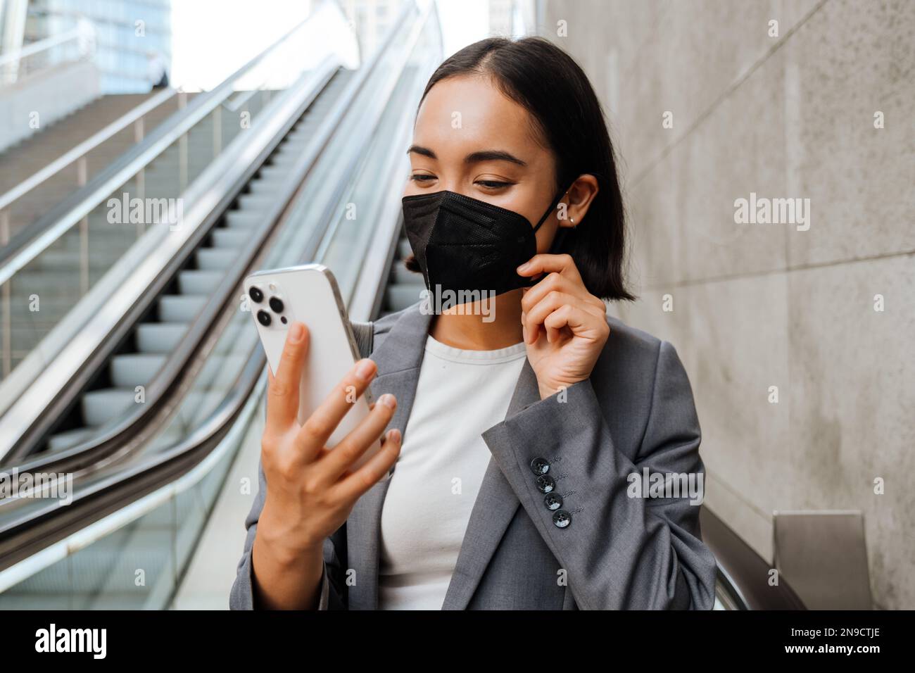 Young asian woman in medical mask looking at mobile phone screen while standing on escalator Stock Photo